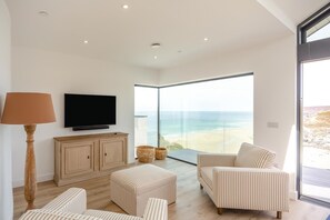 A modern living room with a flat-screen TV, a wooden cabinet, striped ottomans, and a large glass door leading to a balcony with a beach view. - 4 Bed in Porthtowan (oc-45963) (Porthtowan)