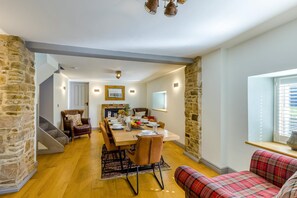 Ein Essbereich mit Holztisch, Stühlen und Kamin. Eine Treppe führt zu einer weiteren Ebene, und ein Sofa mit kariertem Bezug ist vorhanden.