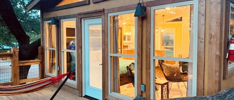 A wooden cabin with large glass doors, a hammock, and a dining area visible through the windows.