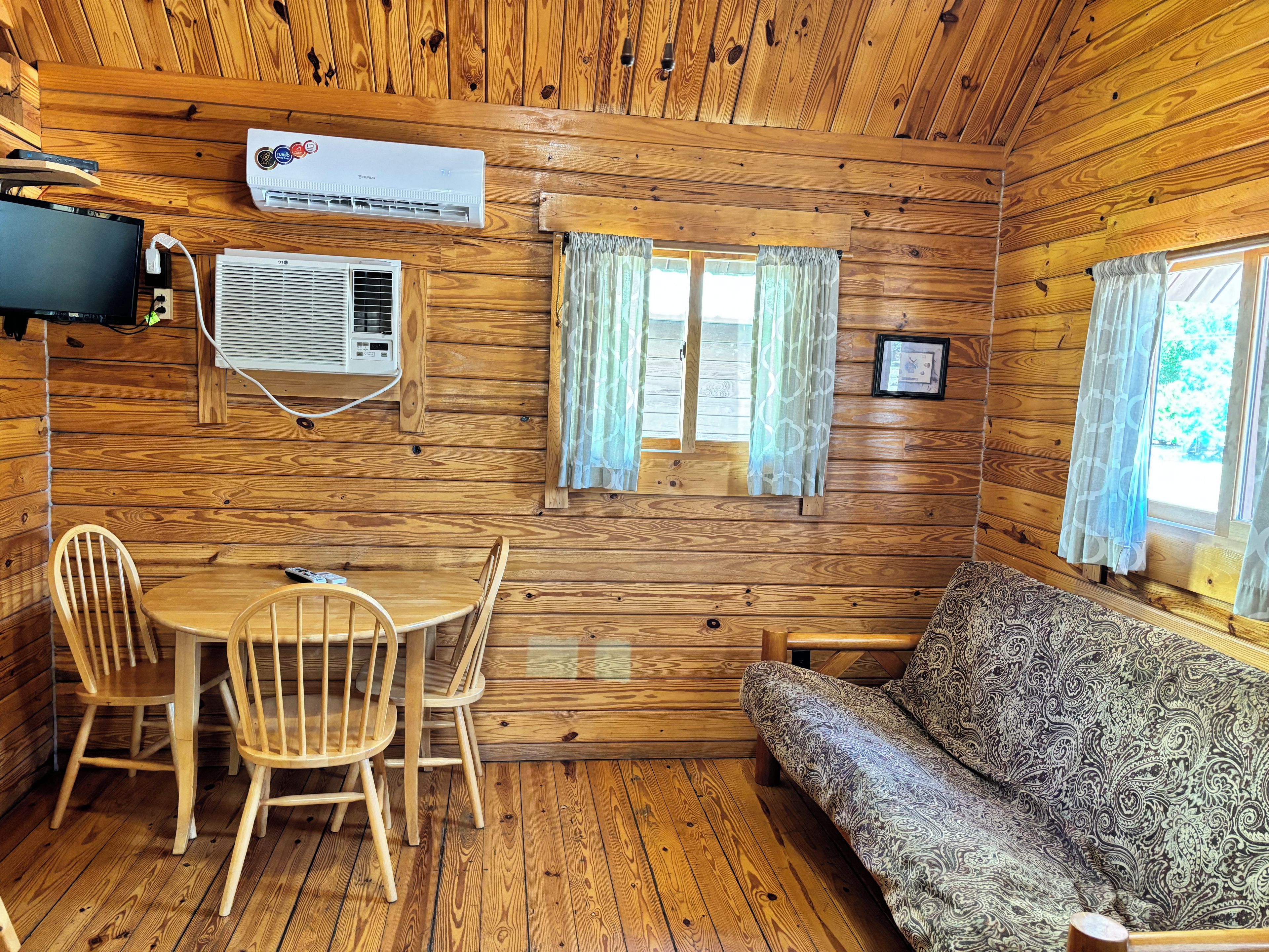 A wooden cabin interior with a dining table, chairs, a sofa, and two windows with curtains.