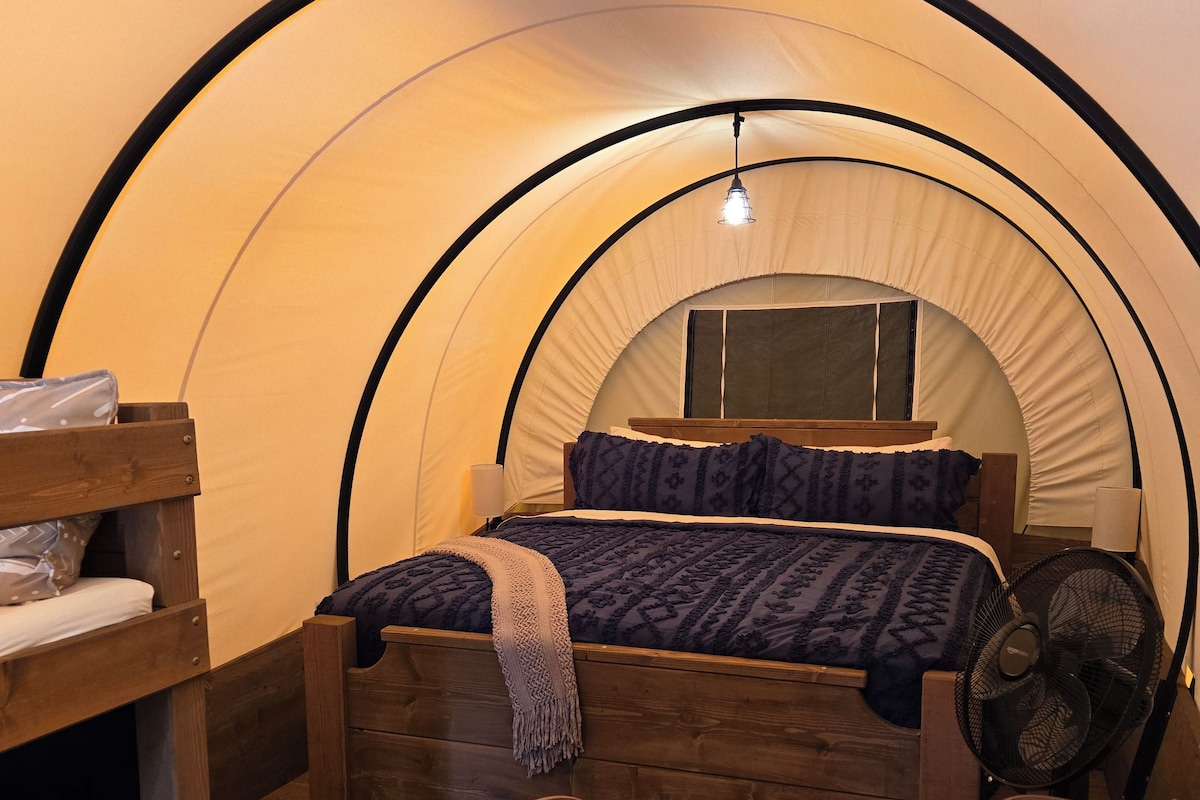 A cozy tent room with a bed, wooden furniture, a table with a glass top, and a fan.