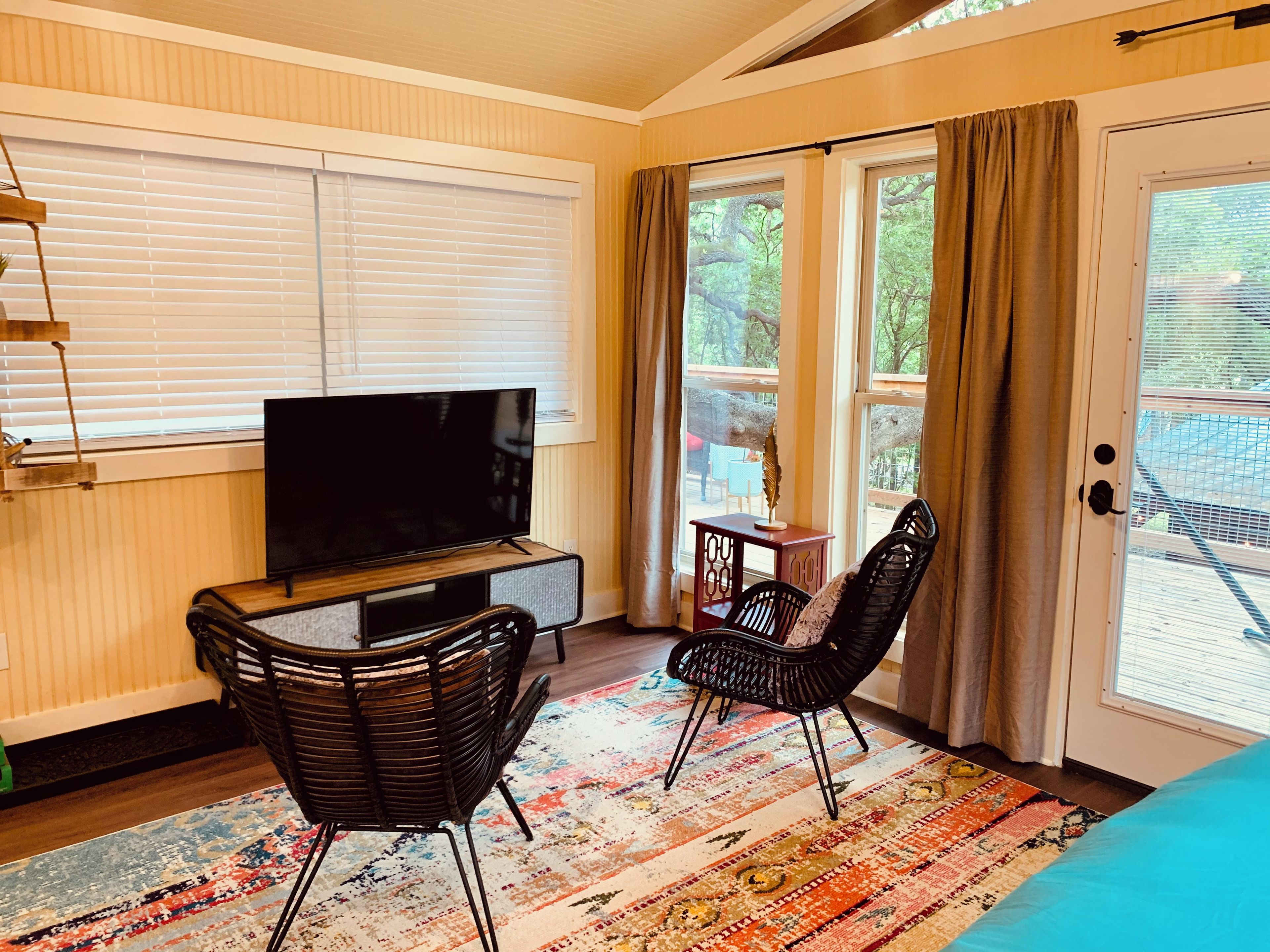 A room with a television on a wooden stand, two black wicker chairs, a colorful rug, and a sliding glass door leading to an outdoor area.