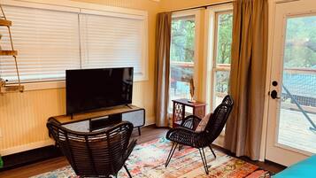 A room with a television on a wooden stand, two black wicker chairs, a colorful rug, and a sliding glass door leading to an outdoor area.