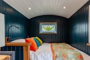 A bedroom with a curved ceiling, a bed with colorful pillows, a window with a floral design, and a nightstand with a lamp. - 1 Bed in Batheaston (oc-f45918) (Batheaston)