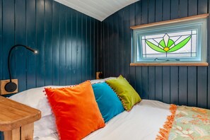 A bedroom with a bed, colorful pillows, a bedside table, a lamp, and a window with a stained glass design. - 1 Bed in Batheaston (oc-f45918) (Batheaston)