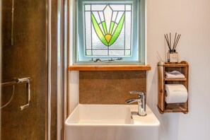 Een badkamer met een witte wastafel, een houten plankje met een flesje kamerdiffuser, en een glas-in-loodraam boven de wastafel.