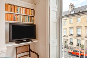 A room with a television on a shelf, a chair, and a view of a street with buildings and a car. - 1 Bed in Bath (oc-r45717) (Bath)