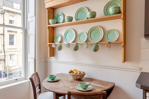 A dining area with a wooden table, chairs, and a wall-mounted shelf with plates and cups. - 1 Bed in Bath (oc-r45717) (Bath)