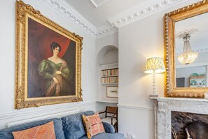 A room with a large framed painting, a bookshelf, a blue sofa, a lamp, and a fireplace. - 1 Bed in Bath (oc-r45717) (Bath)