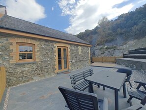 A stone building with a door and window, outdoor seating with benches and a table, and a hilly landscape in the background. - 3 Bed in Talybont (oc-b46034) (Llandre)