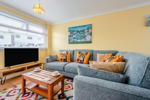Ein Wohnzimmer mit grauem Sofa, bunten Kissen, einem Flachbildfernseher, einem Holztisch und einem Gemälde an der Wand.
