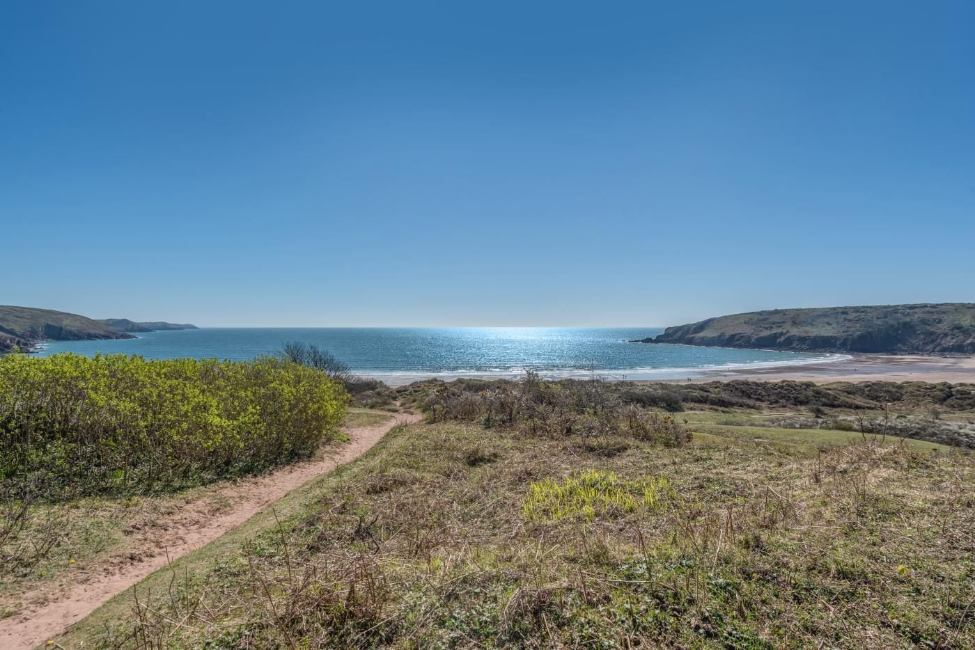 A coastal landscape with a clear sky, a dirt path leading to the beach, and distant hills.