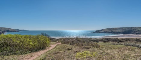 A coastal landscape with a clear sky, a dirt path leading to the beach, and distant hills.