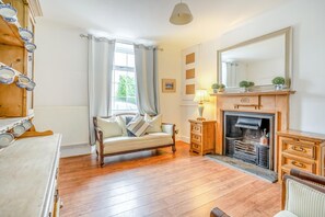 Ein gemütliches Zimmer mit Holzboden, einer Couch, einem Kamin, einem Spiegel und einem Fenster mit Vorhängen.
