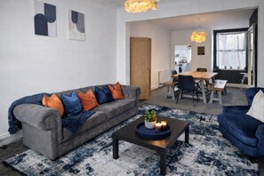 Ein modernes Wohnzimmer mit einem grauen Sofa, blauen und orangefarbenen Kissen, einem Couchtisch mit Kerzen und einem blauen Sessel.