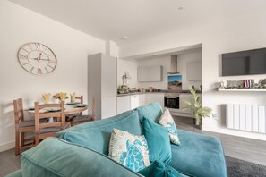 A modern living room with a teal sofa, a dining area with wooden chairs, a kitchen with white cabinets, and a wall-mounted clock. - 2 Bed in Bournemouth (oc-s45959) (Christchurch)
