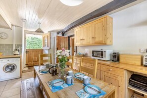 A kitchen with wooden cabinets, a microwave, a kettle, and a dining table set with plates and cutlery. - 3 Bed in Bridport (oc-f45448) (Bothenhampton)
