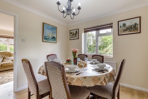Ein Esszimmer mit einem runden Tisch, gedeckt für vier Personen, umgeben von vier Stühlen. An den Wänden befinden sich Gemälde, eine Kristallleuchter und ein Fenster mit Blick auf Grünflächen.
