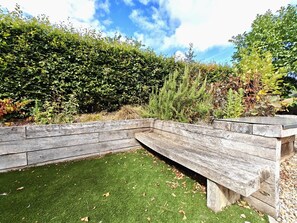 A wooden bench with a built-in planter, surrounded by a well-maintained lawn and dense shrubbery. - 3 Bed in Thornham (oc-h45956) (Thornham)