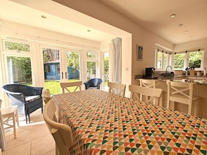 A dining area with a table and chairs, a kitchen with a sink and stove, and a view of a garden through large windows. - 3 Bed in Thornham (oc-h45956) (Thornham)