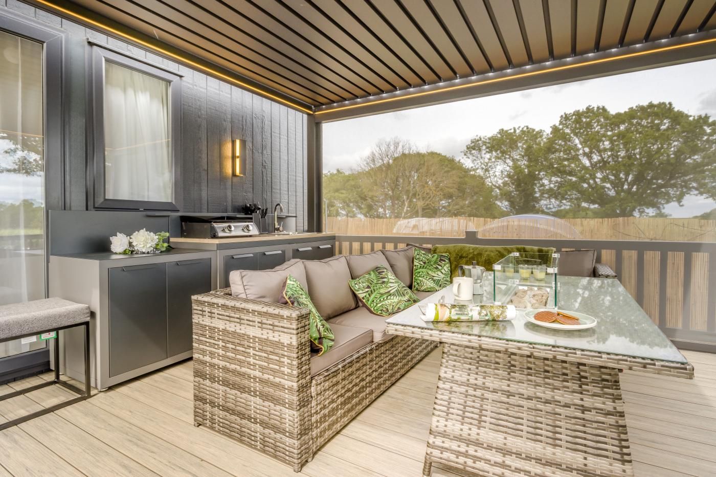 Une cuisine extérieure moderne dotée d’un barbecue intégré, d’un coin salon avec un canapé confortable et des meubles en osier, ainsi que d’une table en verre pour les repas en plein air.