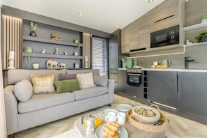 A modern living room with a grey sofa, a kitchen area with a microwave and oven, and a dining table set with breakfast items.