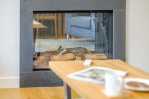 A fireplace with logs, a wooden table with a white mug, and a blue chair in the background.