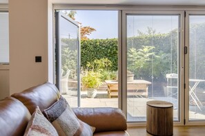 En moderne stue med en lædersofa, et rundt træsidebord og en skydedør, der fører til en udendørs patio med et bord og stole.