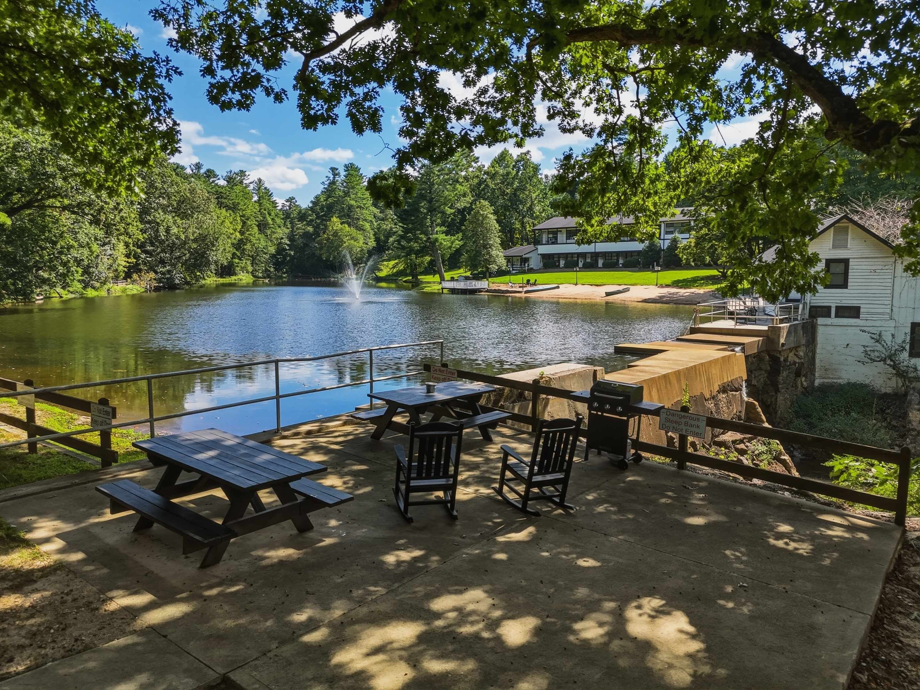 A lakeside picnic area with tables and benches, a wooden deck, and a view of a lake with a fountain.