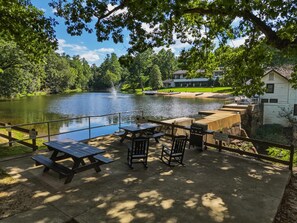 A lakeside picnic area with tables and benches, a wooden deck, and a view of a lake with a fountain.