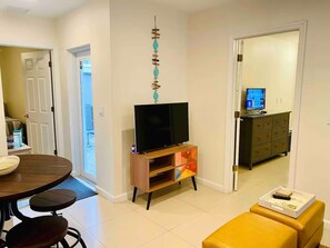 Ein Wohnzimmer mit Fernseher auf einem Holzständer, einem Esstisch mit Stühlen und einem gelben Hocker.