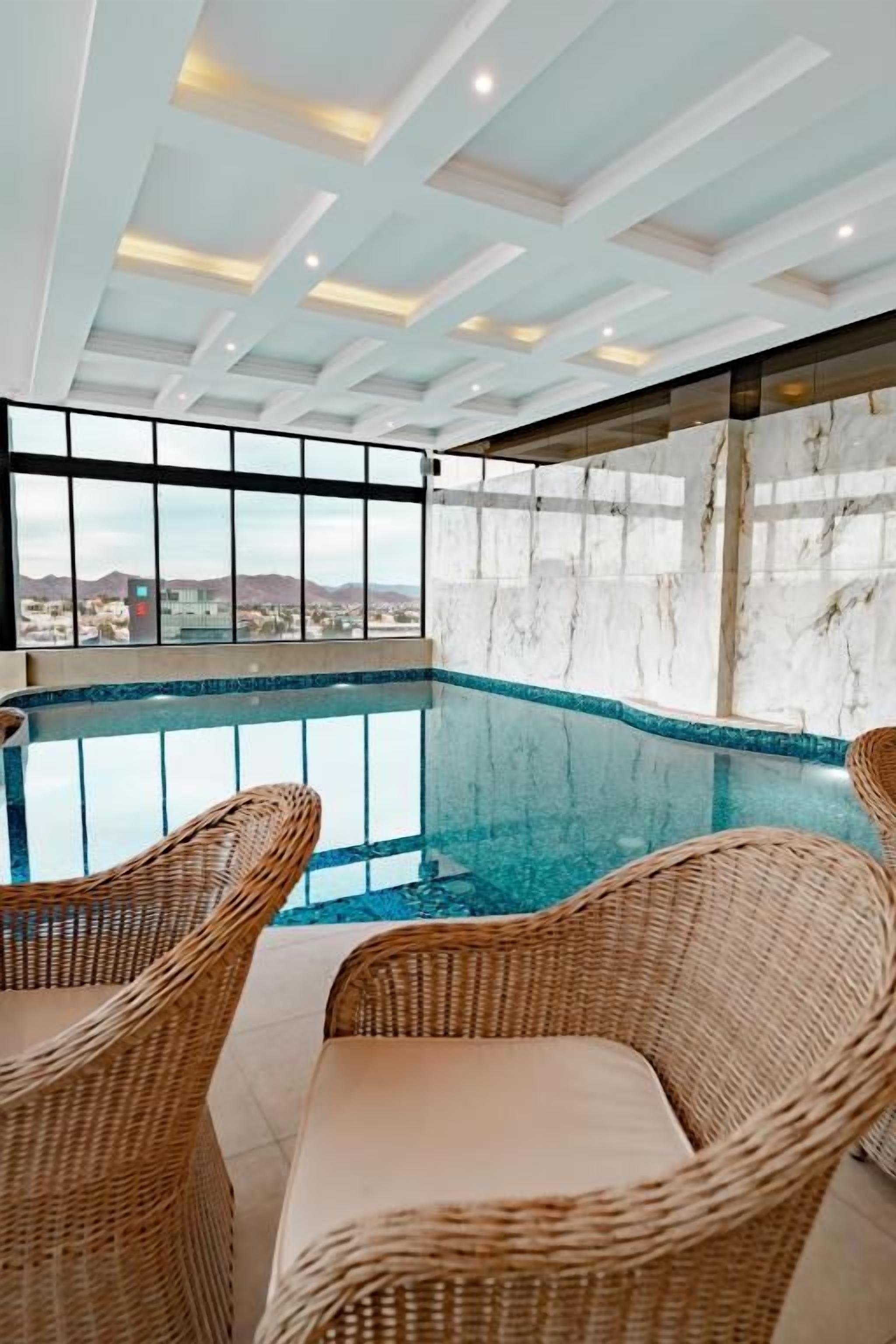 An indoor swimming pool with wicker chairs, a view of the city, and a marble wall.