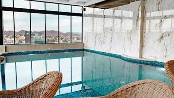 An indoor swimming pool with wicker chairs, a view of the city, and a marble wall.