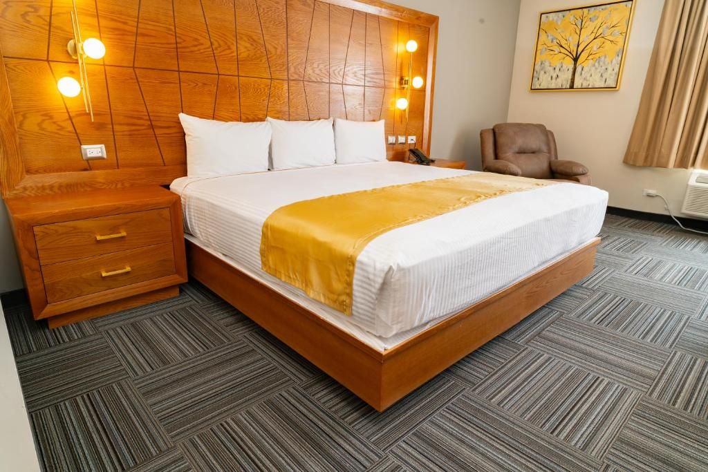Una habitación de hotel con cabecera de madera, cama con ropa blanca y un plaid amarillo, mesita de noche con cajones, un sillón marrón y un cuadro en la pared.