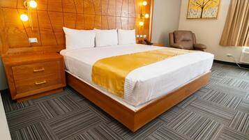 Una habitación de hotel con cabecera de madera, cama con ropa blanca y un plaid amarillo, mesita de noche con cajones, un sillón marrón y un cuadro en la pared.