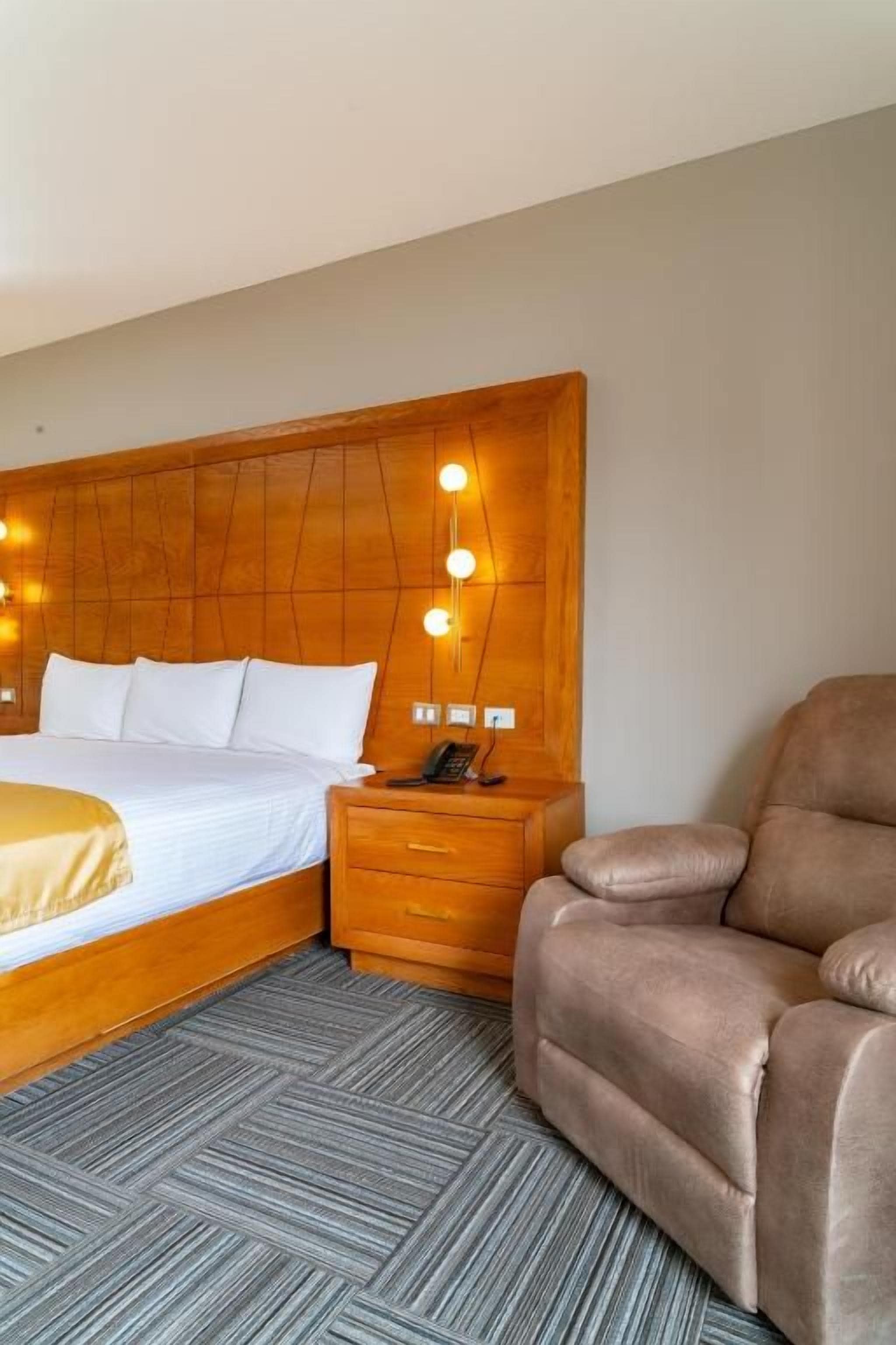 A hotel room with a bed, bedside table, lamp, and a brown armchair.
