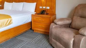 A hotel room with a bed, bedside table, lamp, and a brown armchair.