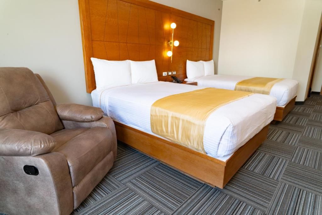 A hotel room with two beds, a sofa, and a wooden headboard with wall-mounted lights.