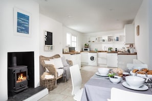 A modern kitchen with white appliances, a dining area with a table set for two, and a fireplace with a visible fire. - 2 Bed in Mousehole (oc-b584390) (Mousehole)