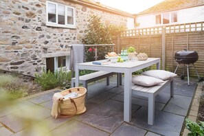A stone house with a patio, a table with cushions, a basket, and a barbecue grill. - 2 Bed in Mousehole (oc-b584390) (Mousehole)