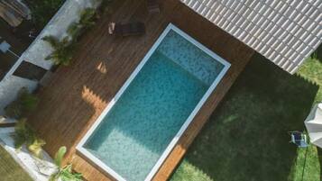 Ein rechteckiger Pool mit einer umlaufenden Holzterrasse, einem Ziegeldach darüber und einem kleinen Gartenbereich mit Pflanzen und einer Bank.