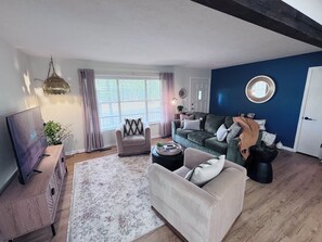 Ein Wohnzimmer mit Sofa, Sessel, Couchtisch, Fernseher und einem großen Fenster mit Vorhängen.