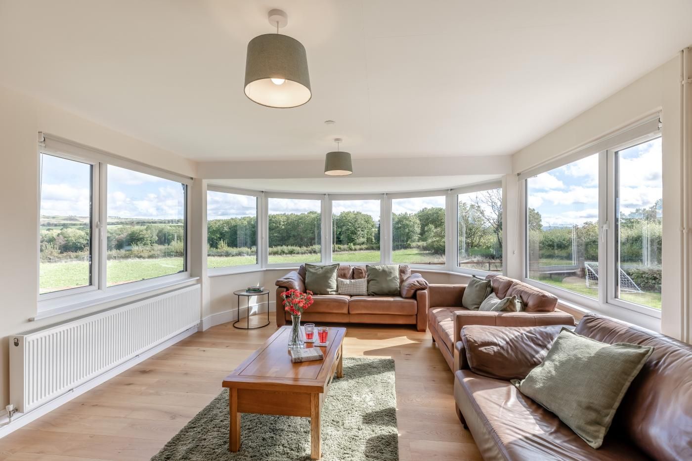 A spacious living room with a large window, a brown leather sofa, a wooden coffee table, and a hanging light fixture.