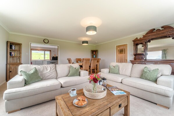 A living room with a sofa set, a coffee table with croissants, and a dining area in the background.