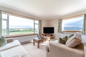 A living room with a television, a coffee table, and a sofa. There are large windows with curtains, and a view of the countryside.
