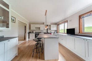 A modern kitchen with white cabinetry, a central island with bar stools, a large window with a view of the outdoors, and a flat-screen TV.