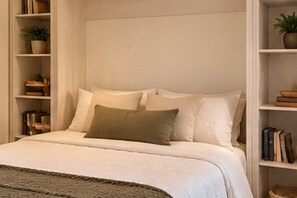 Una cama bien hecha con edredón blanco y varios almohadones, flanqueada por estanterías con libros y una planta en maceta.