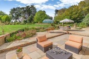 Eine Terrasse mit Korbmöbeln, einem dekorativen Kerzenleuchter und einem Garten mit verschiedenen Pflanzen und Bäumen.