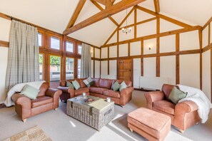 A spacious living room with a high ceiling and exposed wooden beams, furnished with leather sofas and a central wicker coffee table. - 4 Bed in Rhayader (oc-c46317) (Rhayader)