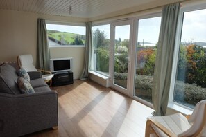 Ein Wohnzimmer mit Sofa, Fernseher und Sessel, mit großen Fenstern und Blick auf Grünflächen und Hügel.